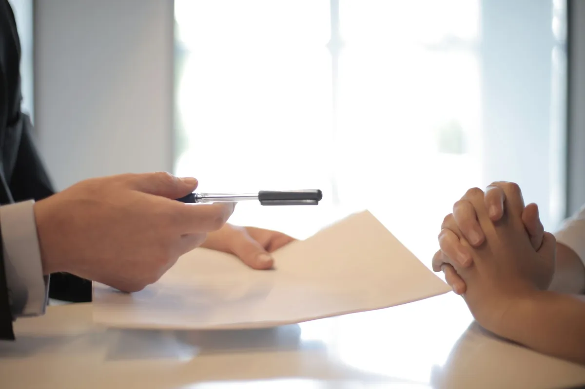 Side view of a faceless formal man handing a pen and paper to a focused woman with clenched hands at a meeting table.