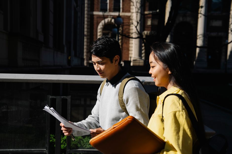 Two students walking with backpacks and papers on a bright day in the city.
